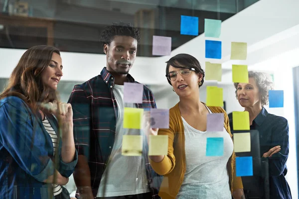 Multiplicando su éxito dividiendo sus tareas. Fotografía de un grupo de colegas que trabajan con notas adhesivas en una pared de vidrio en la oficina. — Foto de Stock