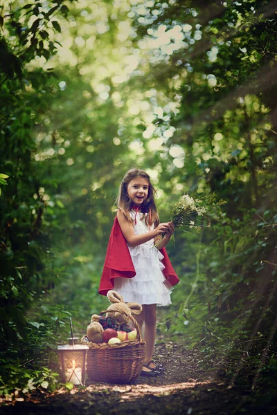 Explorer les merveilles des bois. Portrait d'une petite fille vêtue d'une cape rouge tenant des fleurs dans les bois. — Photo