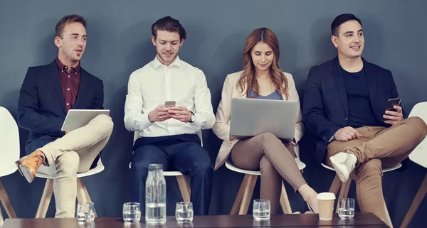 En sus dispositivos inalámbricos mientras esperan. Fotografía de un grupo de empresarios que utilizan diferentes dispositivos inalámbricos mientras esperan una entrevista. —  Fotos de Stock