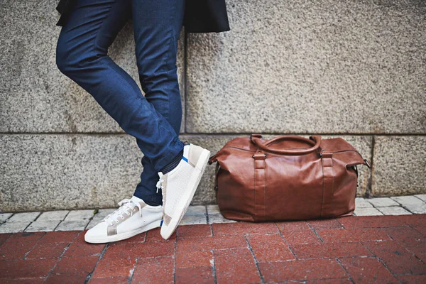 Homem moderno - ir saco ou ir para casa. Tiro cortado de um homem usando jeans e tênis de pé ao lado de sua bolsa em uma calçada da cidade. — Fotografia de Stock