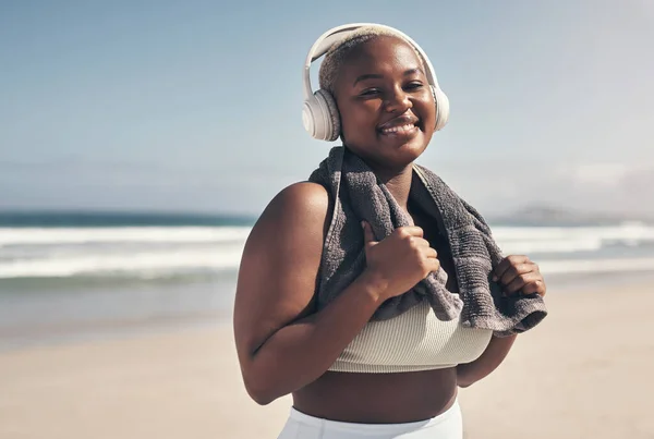 Im working on me, for me. Shot of a sporty young woman wearing headphones and a towel around her neck on the beach. — Stock Photo, Image