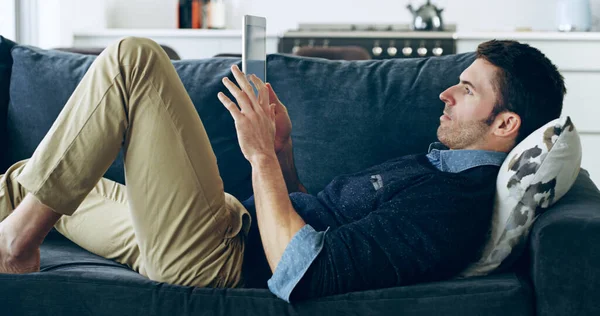 Ein wenig Zeit online verbringen. Schnappschuss eines gutaussehenden jungen Mannes mit einem digitalen Tablet, während er zu Hause auf dem Sofa liegt. — Stockfoto
