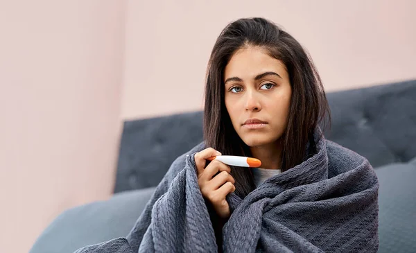 Its official, Ive got the flu. Shot of a young woman taking her temperature while recovering from an illness in bed at home. — Stock Photo, Image