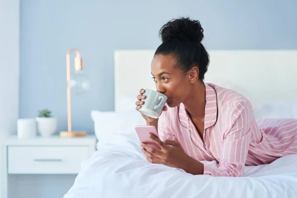 Rien n'a meilleur goût que les commérages matinaux. Tourné d'une jolie jeune femme buvant du café tout en utilisant son téléphone portable dans son lit à la maison. — Photo