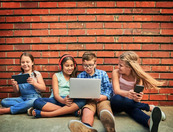 Compartir una infancia despreocupada y conectada. Fotografía de un grupo de niños pequeños usando dispositivos digitales contra una pared de ladrillo. —  Fotos de Stock