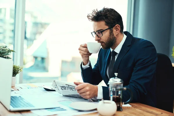 Veamos cómo van los mercados. Tiro de un joven empresario tomando una taza de té mientras lee un periódico en una oficina. — Foto de Stock