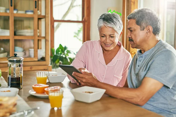 Ich versuche immer noch, das herauszufinden. Aufnahme eines fröhlichen älteren Ehepaares, das gemeinsam auf einem digitalen Tablet surft, während es zu Hause am Tisch frühstückt. — Stockfoto