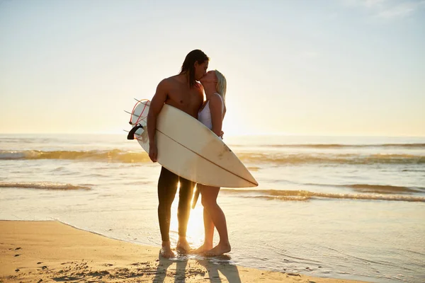 Ich liebe dich, bis dem Meer das Wasser ausgeht. Aufnahme eines liebevollen jungen Paares mit einem Surfbrett, das sich bei Sonnenuntergang am Strand küsst. — Stockfoto