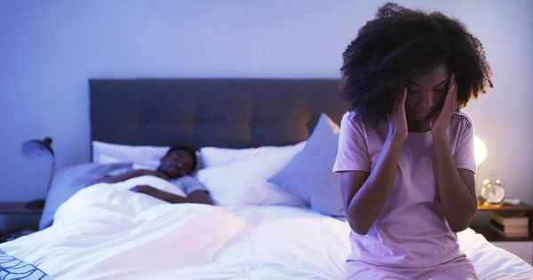 This headache has kept me up the whole night. Shot of a concerned looking young woman sitting on a bed with her husband sleeping in the background. — Stock Photo, Image