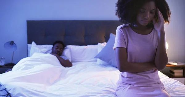 No puedo dormir con este dolor de cabeza palpitante. Fotografía de una joven preocupada sentada en una cama con su marido durmiendo en el fondo. —  Fotos de Stock