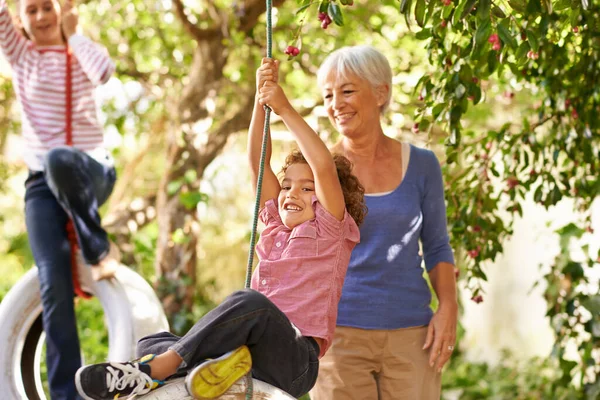 Shot of a woman helping her grandson on a tire swing with her granddaughter in the background.