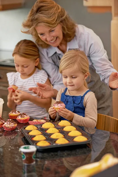 A grandmother and her two grandchildren decorating cupcakes at home.