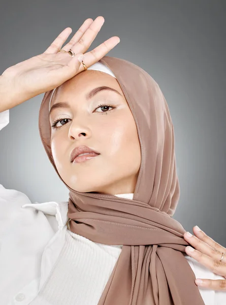 Modest female model wearing makeup with face covered in traditional hijab. Portrait of one beautiful young muslim woman wearing brown headscarf with hand on forehead against grey studio background