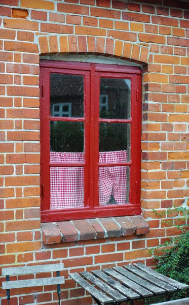 Old window of a brick wall house or home. Ancient red framed casement window on a historic building with a small checkered curtain. Exterior details of a windowsill in a traditional country town.