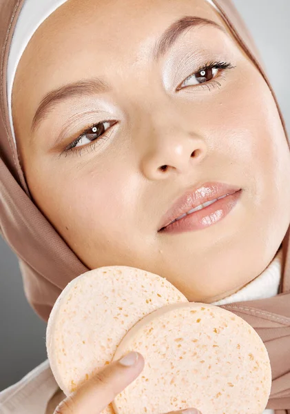 美しいイスラム教徒の女性を保持し 灰色のスタジオの背景に対して彼女の顔に化粧スポンジを使用しています 若い女性はヒジャーブを着用し 彼女の美しさのスキンケアルーチンを行う 健康な皮膚の世話をする — ストック写真