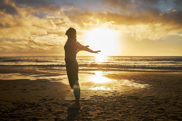 Rear View of a silhouette woman stretching with her arms out at the beach during sunset. Calm relaxed female feeling freed and enjoying a golden sunset out in nature. Golden sunset during dusk.