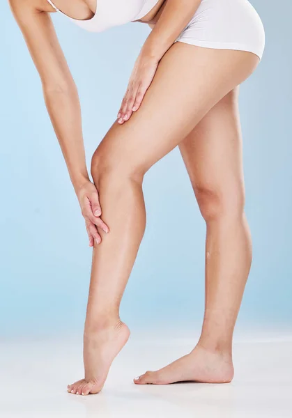 Woman stroking her shaved and smooth legs isolated on a blue studio background. Slim female touching her skin after hair removal, waxing or depilation treatment. Showing her beautiful long legs.