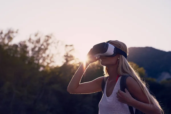 Woman hiking at sunrise using VR goggles to explore the metaverse. Blond woman on holiday hiking in a forest using VR goggles to experience augmented virtual reality.
