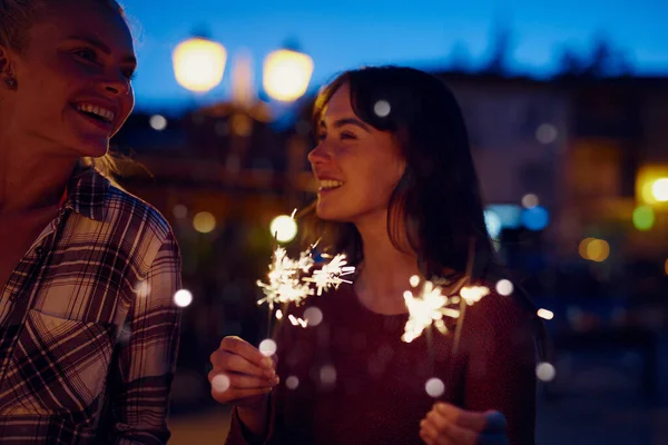 Young woman smiling at her friend and celebrating playing with fireworks. Two friends on holiday together celebrating with fireworks at night.Happy woman on holiday with friend playing with fireworks.