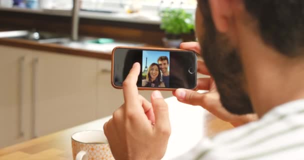 Homem Olhando Através Fotos Seu Celular Enquanto Sentado Sua Cozinha — Vídeo de Stock