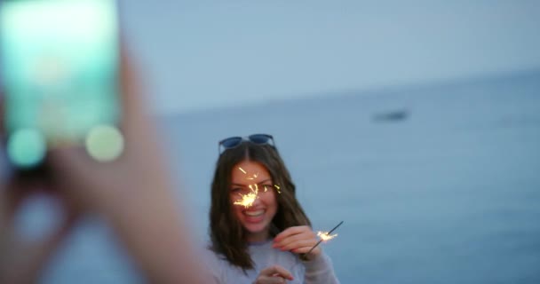 写真を撮られた幸せな女性がビーチでキラキラと遊んでいる 花火で遊ぶ友人の写真を撮る女性 — ストック動画