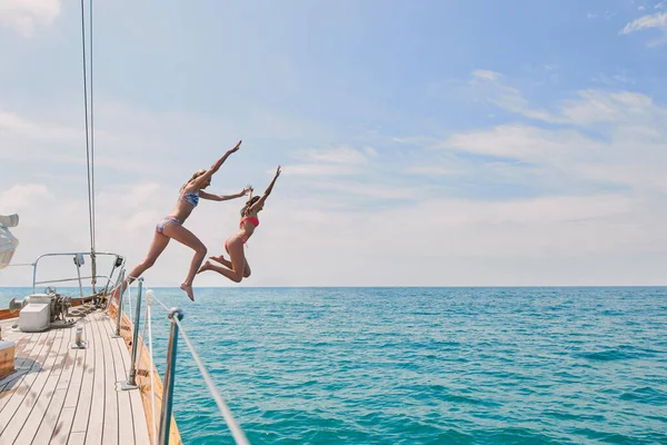 Carefree excited young women jumping from boat to swim in the ocean. Two friends on a holiday cruise together jumping from boat into the ocean to swim. Excited young women jumping off boat together.