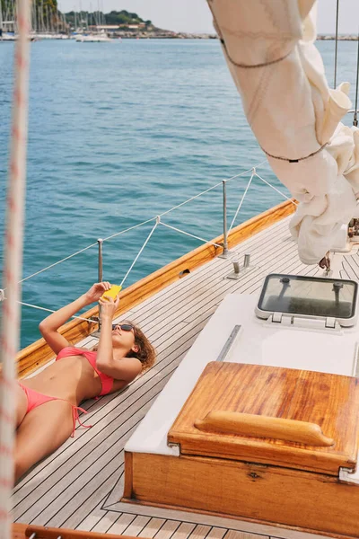 Young woman in a red bikini sunbathing on a boat lying down using her smartphone to send messages. Woman lying on boat using smartphone to scroll apps while sunbathing
