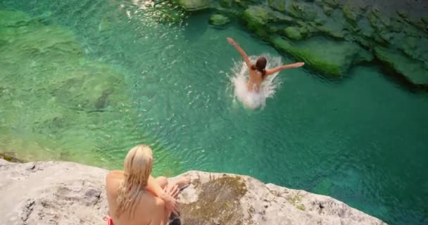 二人の興奮した友人は崖からジャンプした後 湖で泳ぐ 崖からビキニで2人の女性が湖で泳ぐ 休暇中の女性一緒に崖から湖に飛び込むに泳ぐ — ストック動画