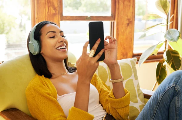 Hispanic woman using her smartphone and listening to music on headphones while comfortable and relaxing in a bright living room. A young female sitting on a chair texting on her cellphone at home.