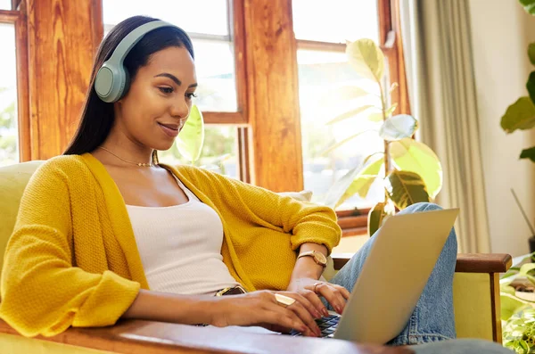Hispanic woman typing on a laptop and listening to music on headphones, smiling and working in a bright living room. A young female sitting on a chair using modern technology at home in lockdown.