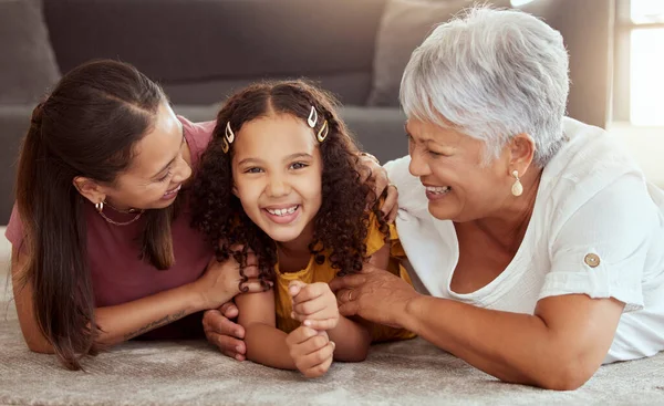 Portrait of mixed race child with single mother and grandmother in home living room. Smiling hispanic girl bonding with parent and senior in lounge. Happy three generations lying together on floor.