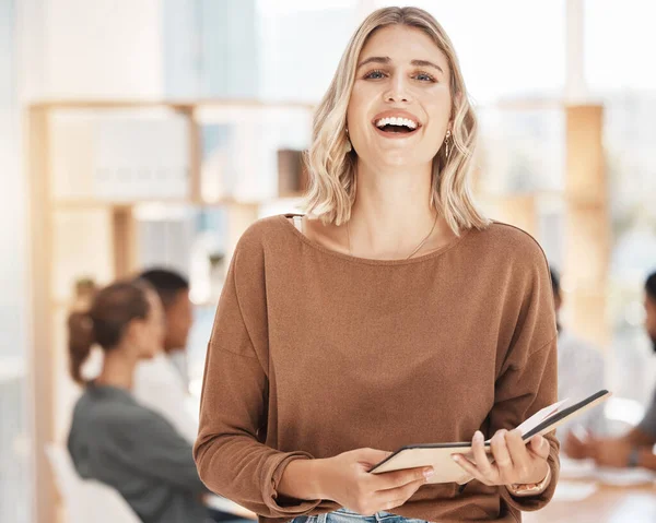 Portrait of a confident young caucasian businesswoman laughing while holding notebook planner in an office with her colleagues in the background. Cheerful ambitious entrepreneur and determined leader