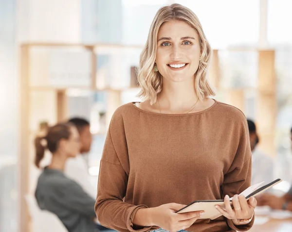 Portrait of a confident young caucasian businesswoman holding notebook planner in an office with her colleagues in the background. Ambitious entrepreneur and determined leader ready for success in a
