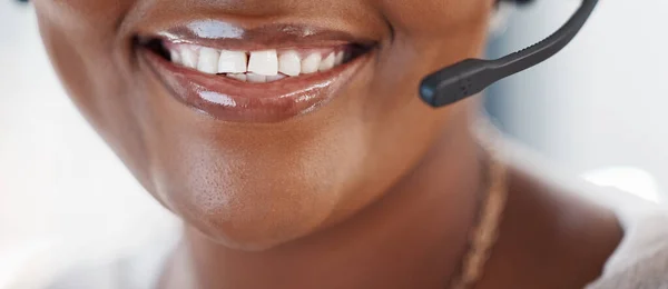 Closeup of one happy african american call centre telemarketing agent with big smile talking on headset while working in office. Face of confident friendly female consultant operating helpdesk for