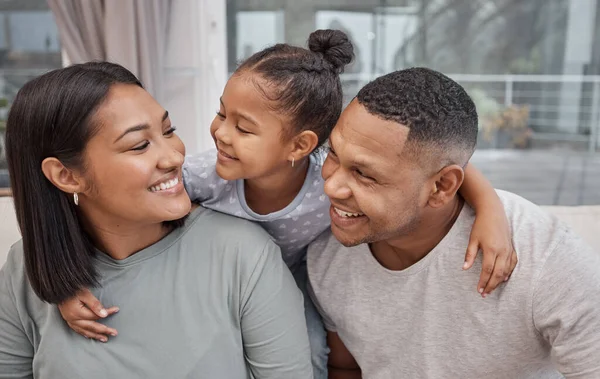 Happy and affectionate young mixed race family of three sitting on a sofa in the living room at home. Married couple sitting with their cute little girl in the lounge. Mom, dad and adorable daughter.