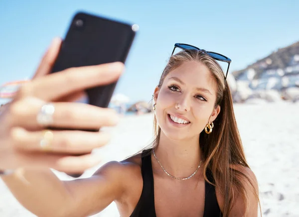 One beautiful young caucasian woman relaxing and taking a selfie on her phone while sitting on the beach. Enjoying a summer vacation or holiday outdoors. Taking time off and getting away from it all.