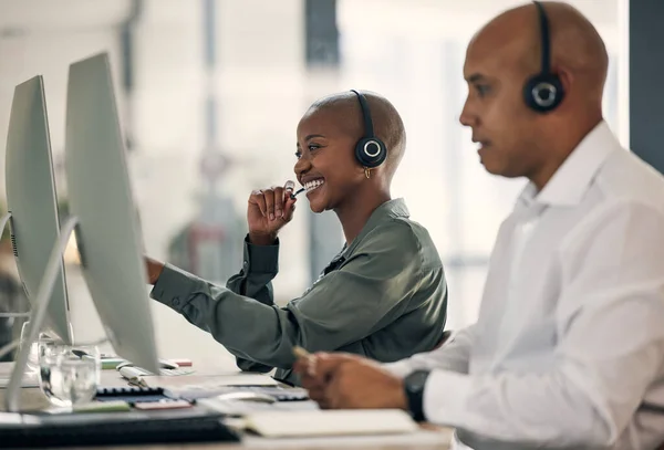 Happy young african american call centre telemarketing agent talking on a headset while working on a computer in an office alongside a colleague. Confident friendly female consultant operating