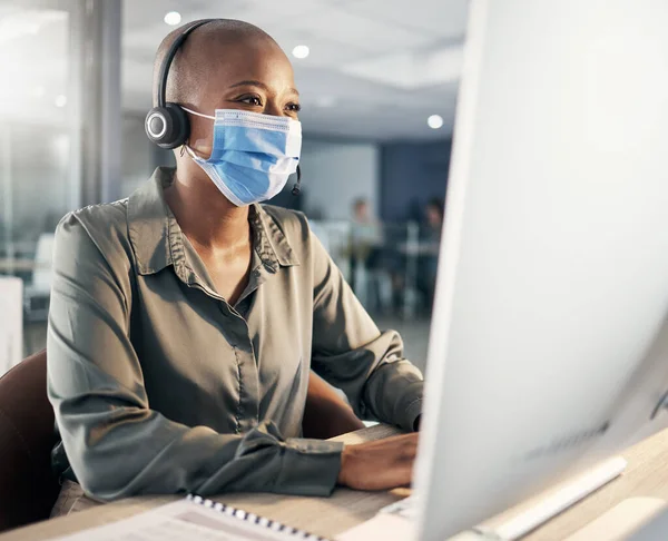 One african american call centre telemarketing agent wearing face mask as health and safety protocol talking on headset while using computer in an office. Female consultant operating helpdesk for