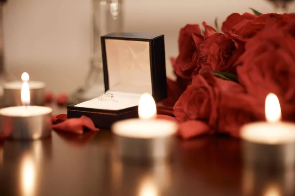 Closeup of romantic engagement ring box, fresh red roses and candles in an empty room at night. Getting ready and prepared for surprise proposal. Loving couple popping the question with sweet gesture.