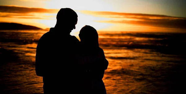 Silhouette affectionate young couple sharing an intimate moment on the beach. Backlit happy husband and wife enjoying a summer day by the sea. They love spending time together on the coast at sunset.