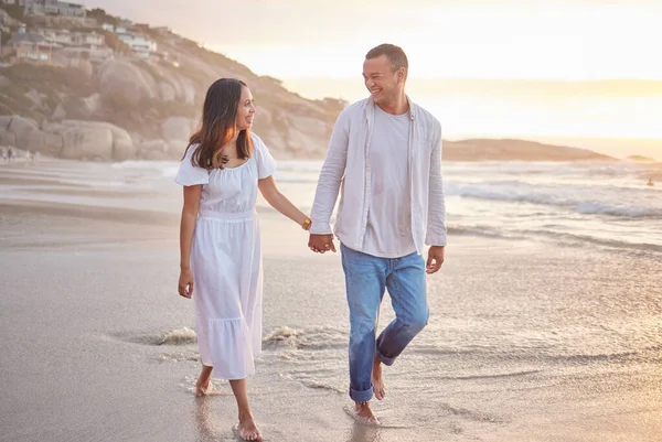 Affectionate mixed race couple holding hands and walking along the beach. Husband and wife hand in hand in the sand by the sea. Enjoying a romantic walk by the seashore at sunset. Love is in the air.