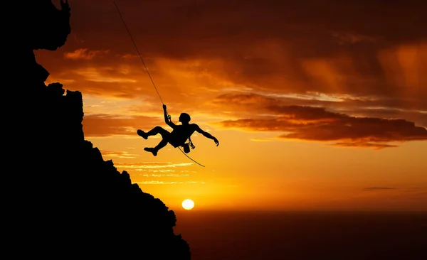 Silhouette of a young male hiker abseiling down the side of a mountain backlit against a sunset sky. Active and sporty man mountain climbing up towards a cliff. Hes a thrill seeking adrenaline junkie.