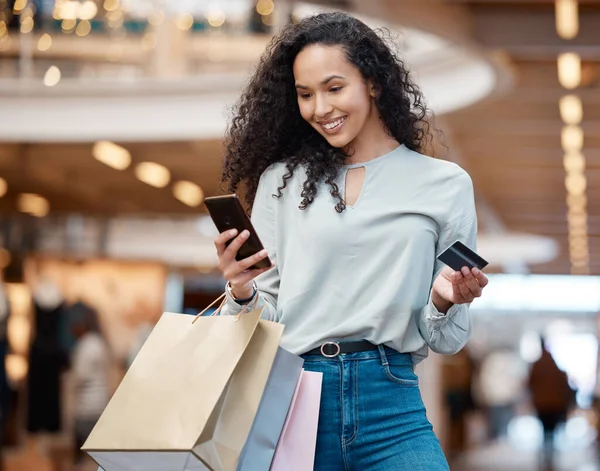 One beautiful mixed race woman standing with her phone and credit card while shopping in a mall. Young hispanic woman carrying bags, spending money, looking for sales and enjoying retail therapy.