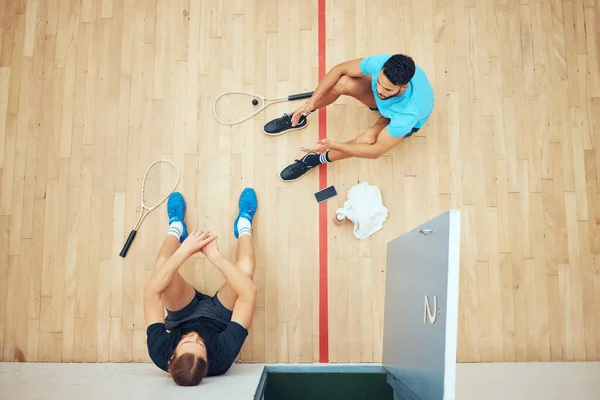 Above view of two unknown athletic squash players sitting together after playing court game. Fit active mixed race and caucasian athletes resting after training practice in sports centre. Sporty men.