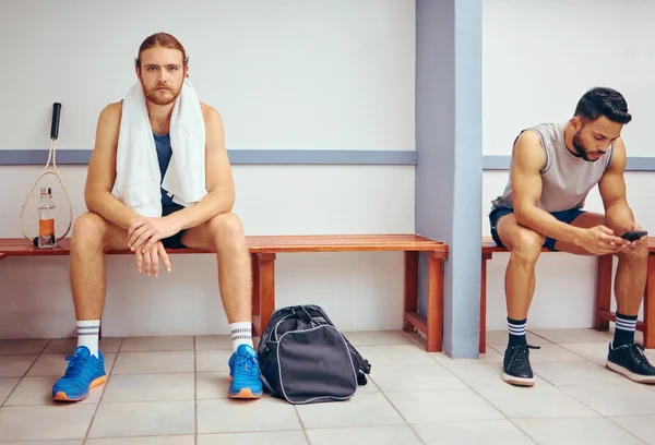 Portrait of a caucasian athlete sitting in a locker room. Two men sitting in a gym locker room together. Serious player relaxing in a gym together. Professional players resting together.