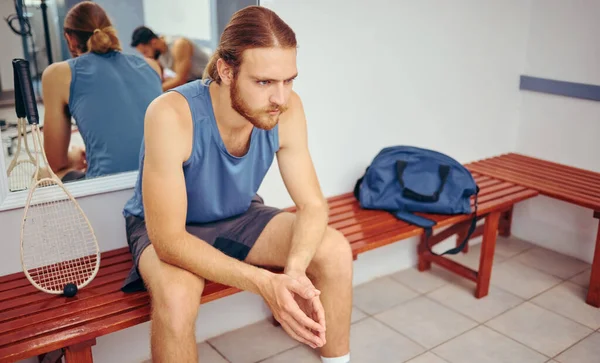 Caucasian player sitting in a locker room with his squash racket. Two players resting together in a gym locker room. Fit young athlete sitting in a locker room thinking about his match.