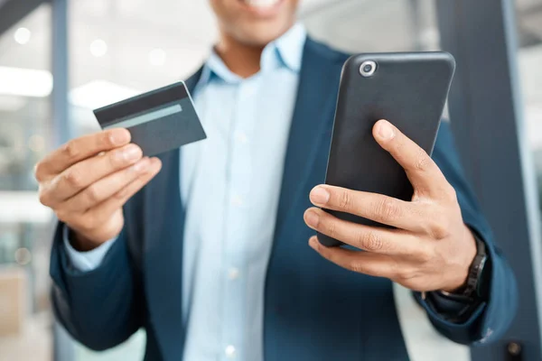 Closeup of a mixed race businessman holding and using a credit card and phone alone at work. One hispanic male businessperson making an online purchase with his debit card and cellphone.