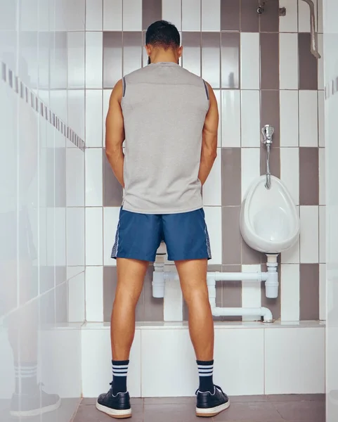 Back of athletic man urinating in a bathroom. Fit man taking a break from his workout to urinate in his gym toilet. Professional squash player taking a break to urinate.