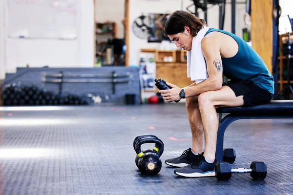 One exhausted young caucasian man taking a rest break to catch his breath and drink water while training in a gym. Fit guy sweating while sitting on a bench with a towel around his neck after an
