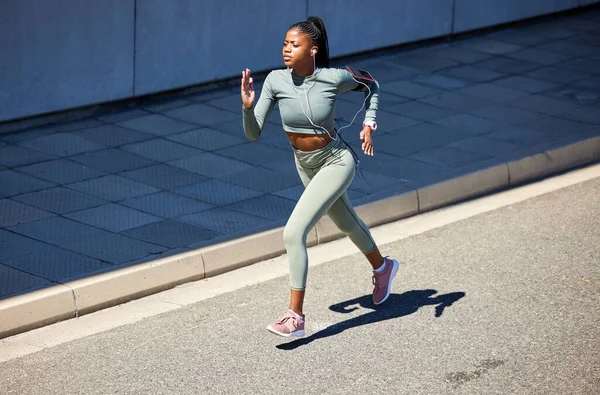 Fit and active African American woman listening to music through earphones and running alone in the city. Black woman focused on fitness and speed while jogging and wearing her phone in an arm holder.